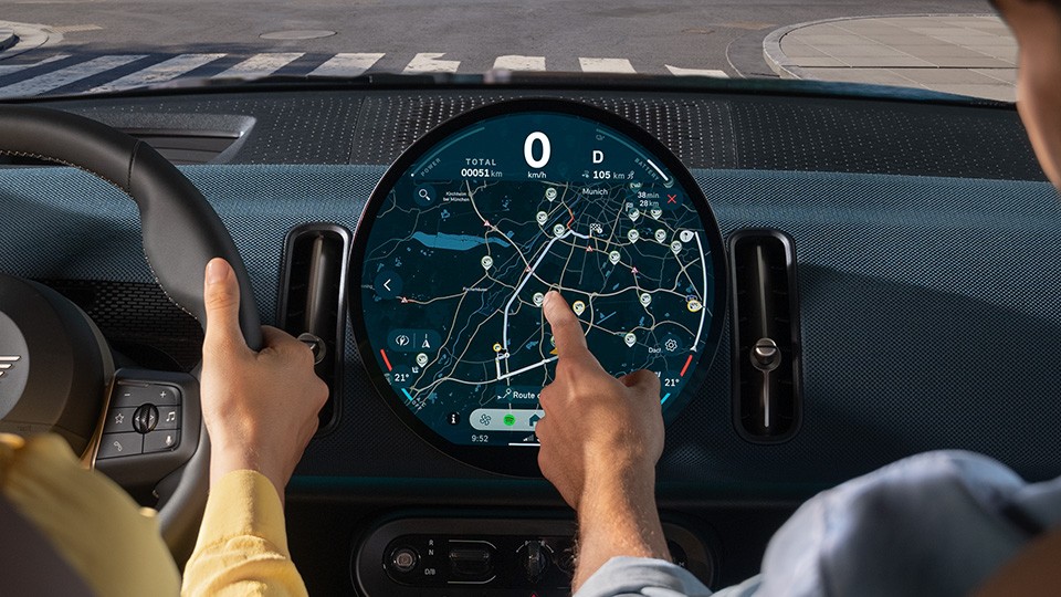 MINI Electromobility - OLED cid with navigation. Driver and passenger interacting with the circular MINI infotainment display showing navigation.